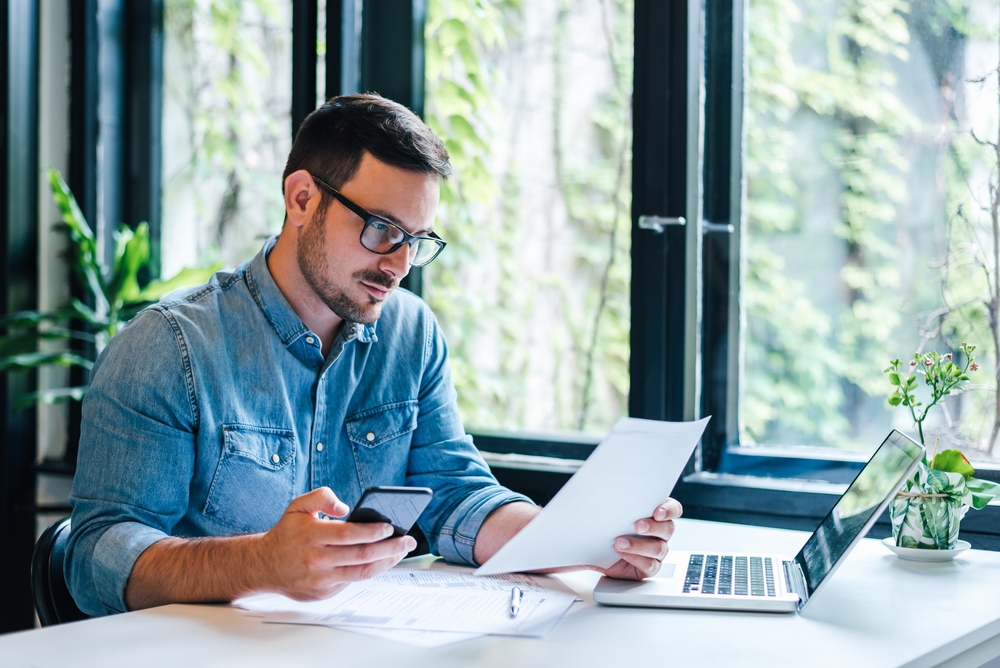 Hombre revisa documentos en smartphone y laptop